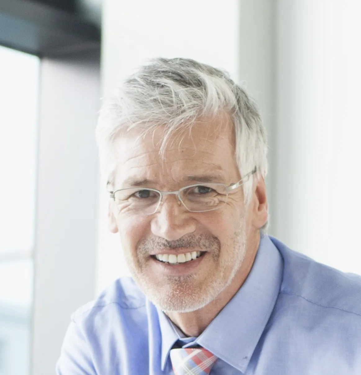 Ältere männliche Führungskraft mit grauem Haar, Brille, blauem Hemd und kariertem Krawatte lächelt.